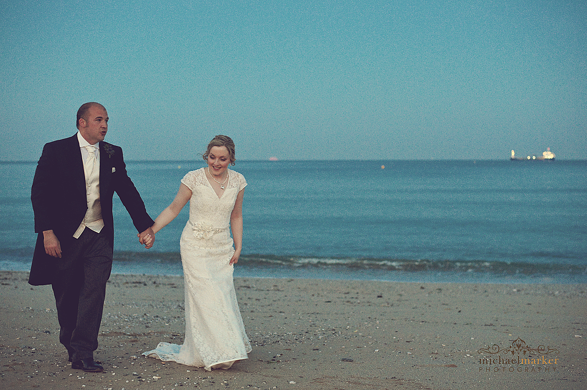 Bride and groom walking hand in hand along Falmouth beach in the early evening