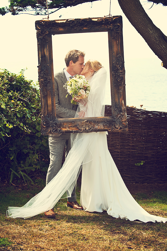 Bride and groom kissing in DIY phtobooth frame hanging from a tree at Polhawn