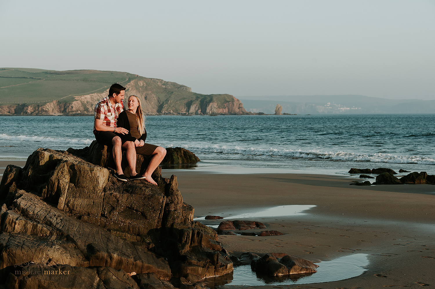 Engaged couple look into each others eyes whilst sitting on rocks at Bigbury beach with Bantham in the background.