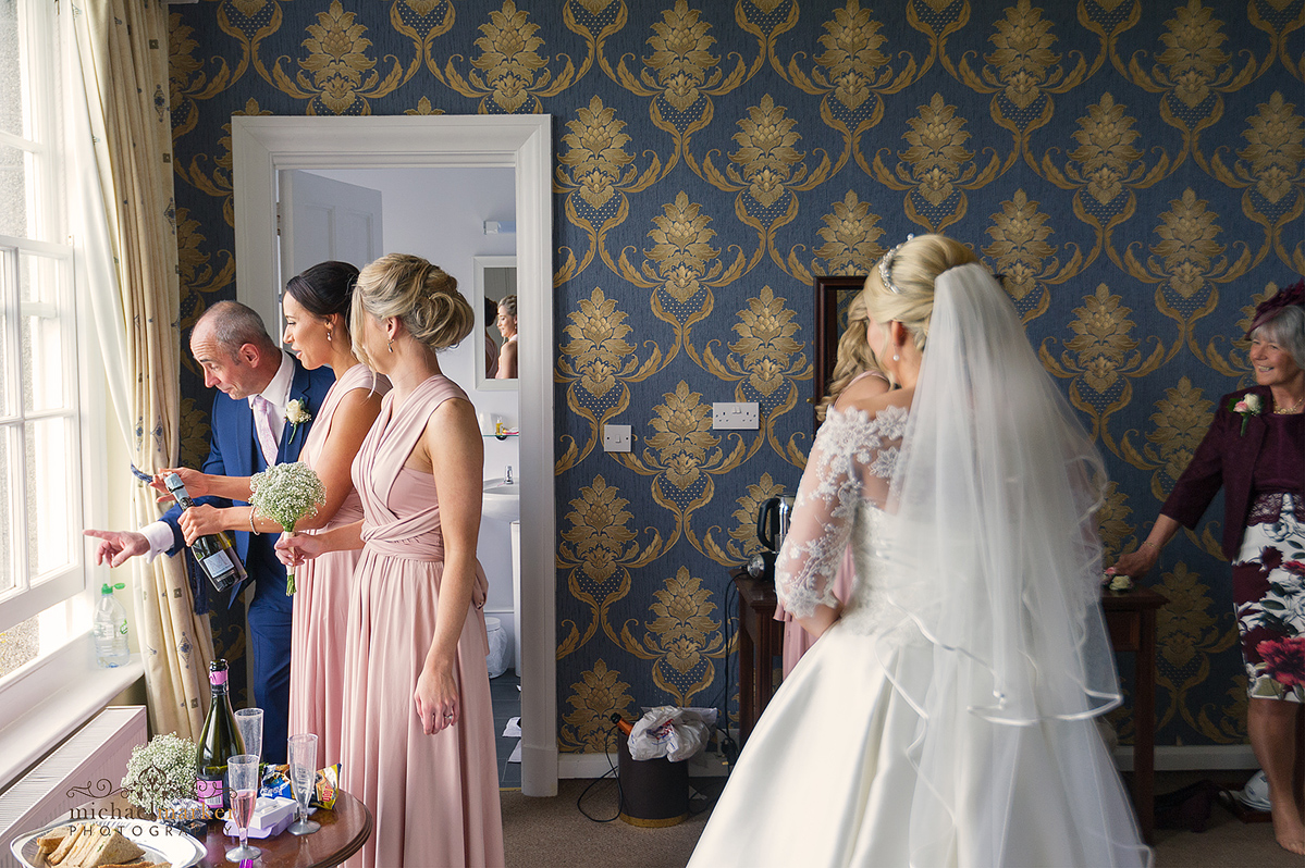 Father and bridesmaids opening bottle of prosecco and looking out window. Bride and mother look on.