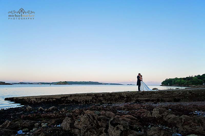 Bride and groom embrace at Plymouth waterfront at dusk.