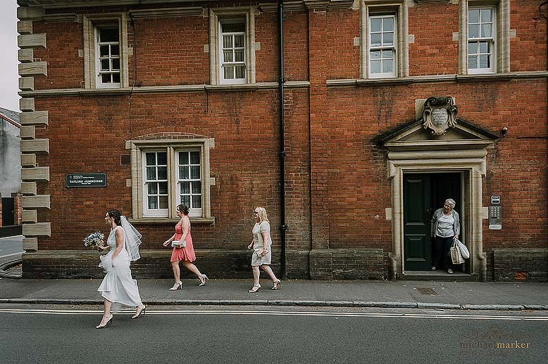 Bride, bridesmaid and mother walk through Salisbury to the wedding ceremony whilst a passerby looks on.