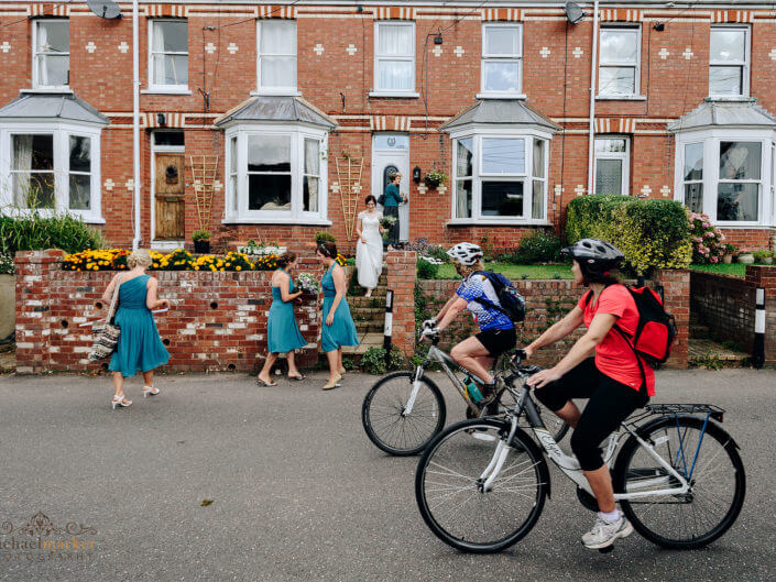 Exeter wedding photographers documentary photo of cyclists riding past bride and bridesmaid as they leave home .
