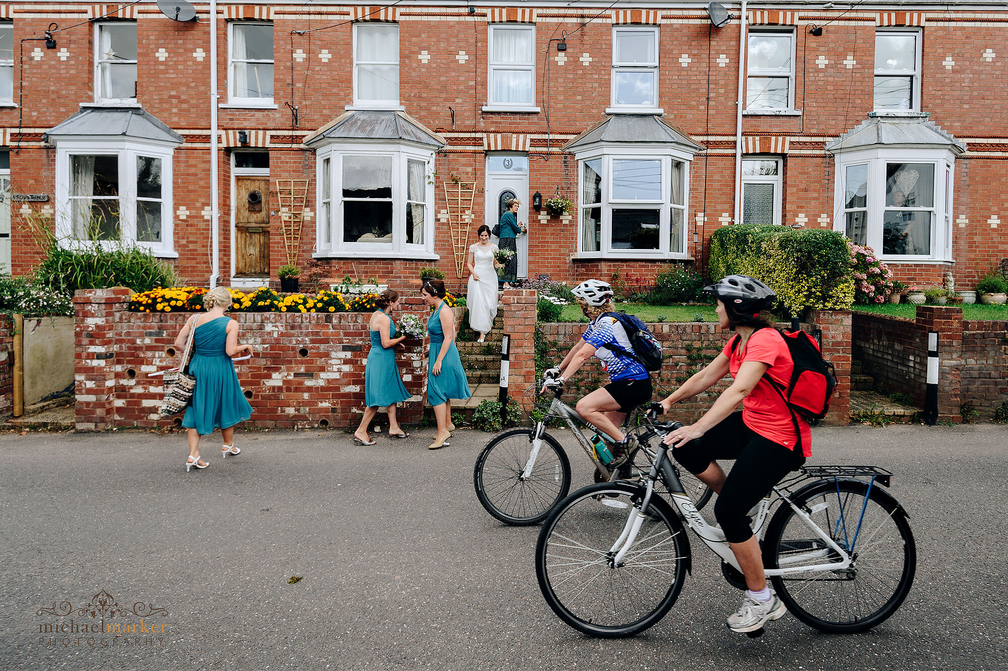 Exeter wedding photographers documentary photo of cyclists riding past bride and bridesmaid as they leave home .