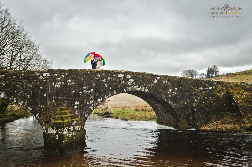 Bride and groom holding colourful umbrellas on Two Bridges Hotel bridge over the river.